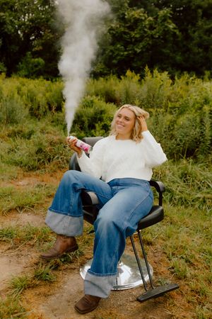 Smiling blonde woman sitting in barber chair outdoors holding pink smoke flare in nature.