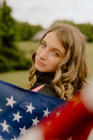 Young blonde woman with curly hair holding American flag outdoors in green field.