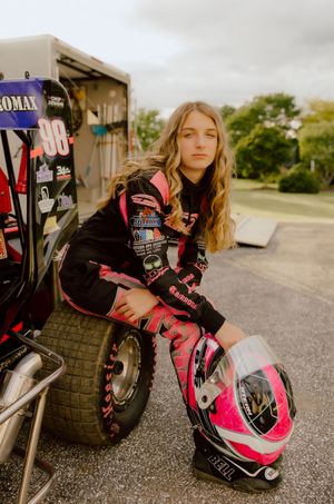 Young female racer in pink and black suit holding helmet beside race car.