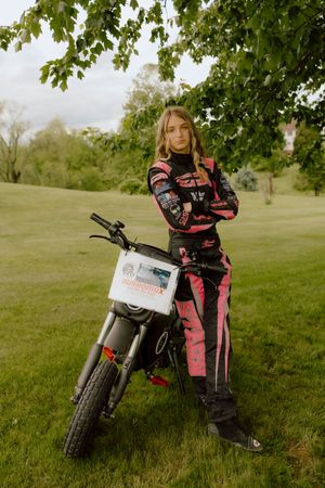 Young woman in pink and black racing suit posing with motorcycle under tree.