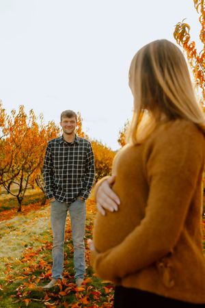 Smiling man in plaid shirt stands in autumn orchard facing pregnant woman.