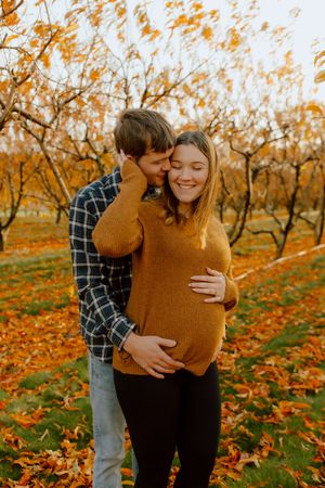 Couple in autumn orchard, man embracing pregnant woman from behind among orange fall leaves.