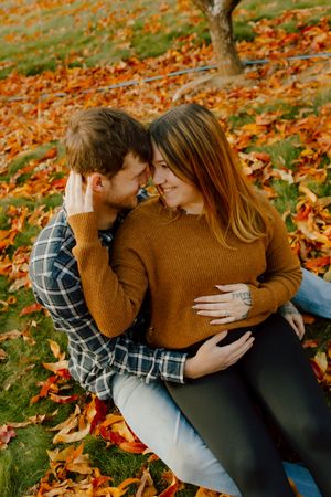 Couple sits together in colorful fallen autumn leaves, sharing an intimate forehead-to-forehead moment.