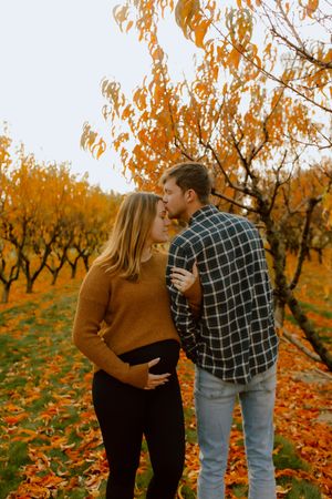 Pregnant couple embraces tenderly among vibrant orange autumn foliage in an orchard setting.
