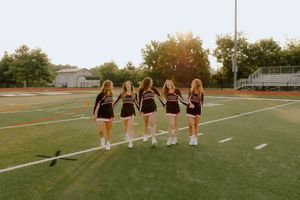 Six cheerleaders in maroon uniforms walk together across a sunny football field at golden hour.