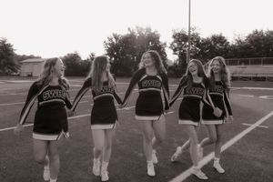 Black and white photo of five cheerleaders in uniforms walking on football field.