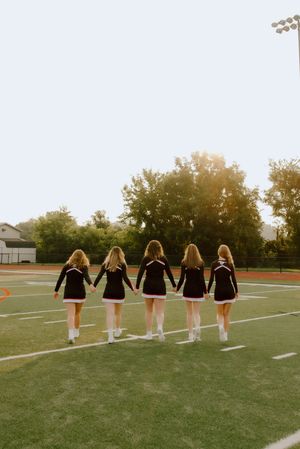 Five cheerleaders in black uniforms holding hands walking across a football field at sunset.