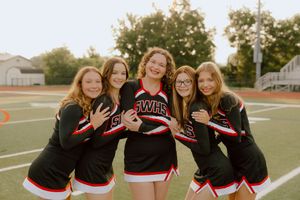 Five cheerleaders in black and red uniforms smiling together on football field.