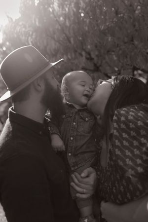 Black and white photo of parents kissing their laughing baby in sunlit outdoor setting.