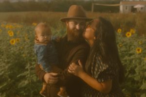 A bearded man in a hat holds a baby while a woman kisses his cheek in a sunflower field.