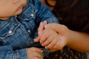 Close-up of mother tenderly holding small baby's hand, child wearing denim jacket.