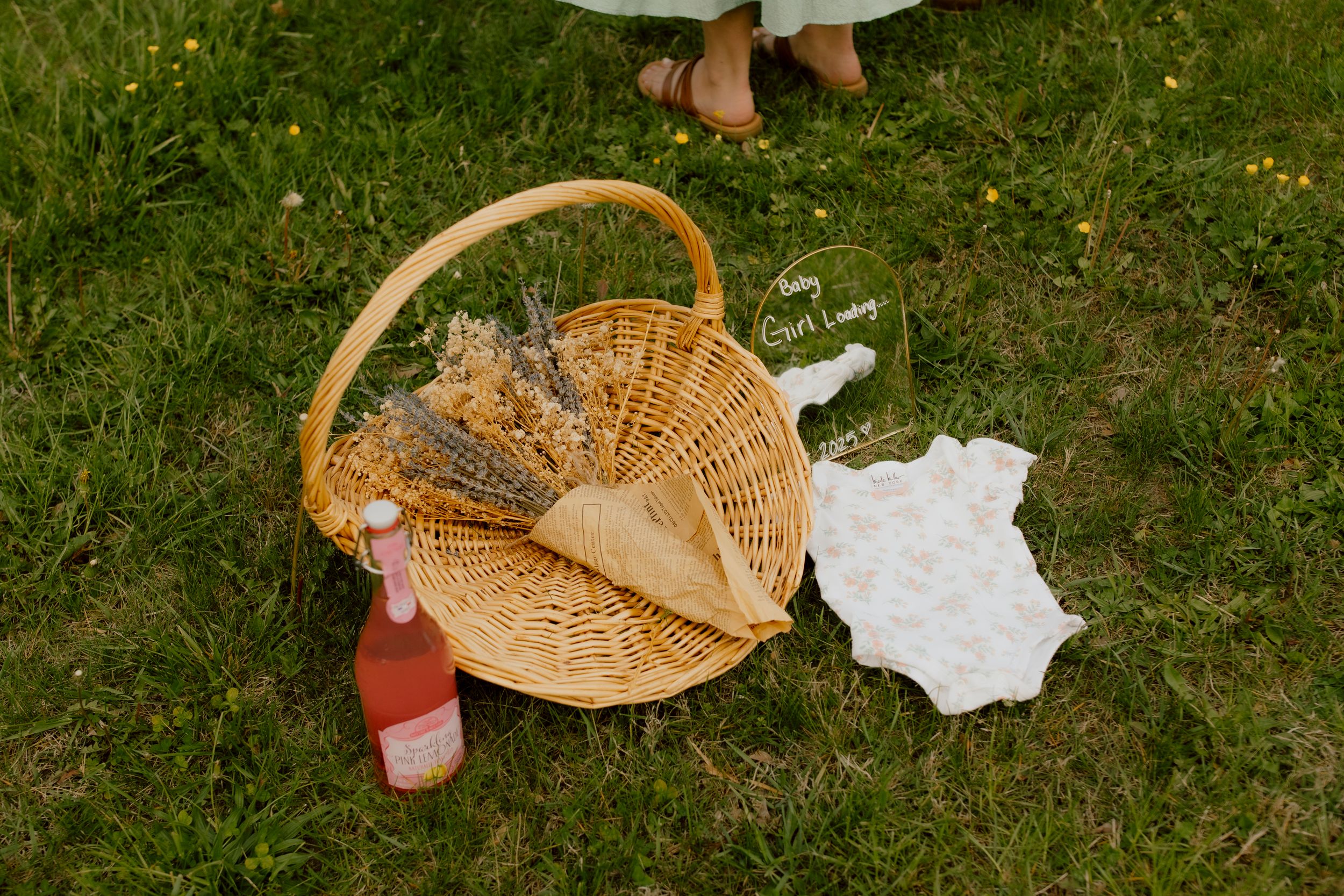 Wicker picnic basket with dried flowers, pink bottle, and white floral baby onesie on green grass.