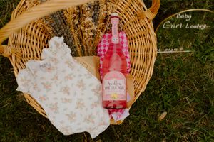 Pink sparkling wine bottle and floral baby outfit in a wicker basket on grass for a baby girl reveal.