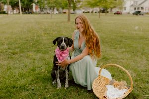 Smiling redhead woman posing with black spotted dog wearing pink bandana in park.