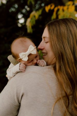 Mother smiles joyfully while holding her infant with a white bow headband against autumn foliage.