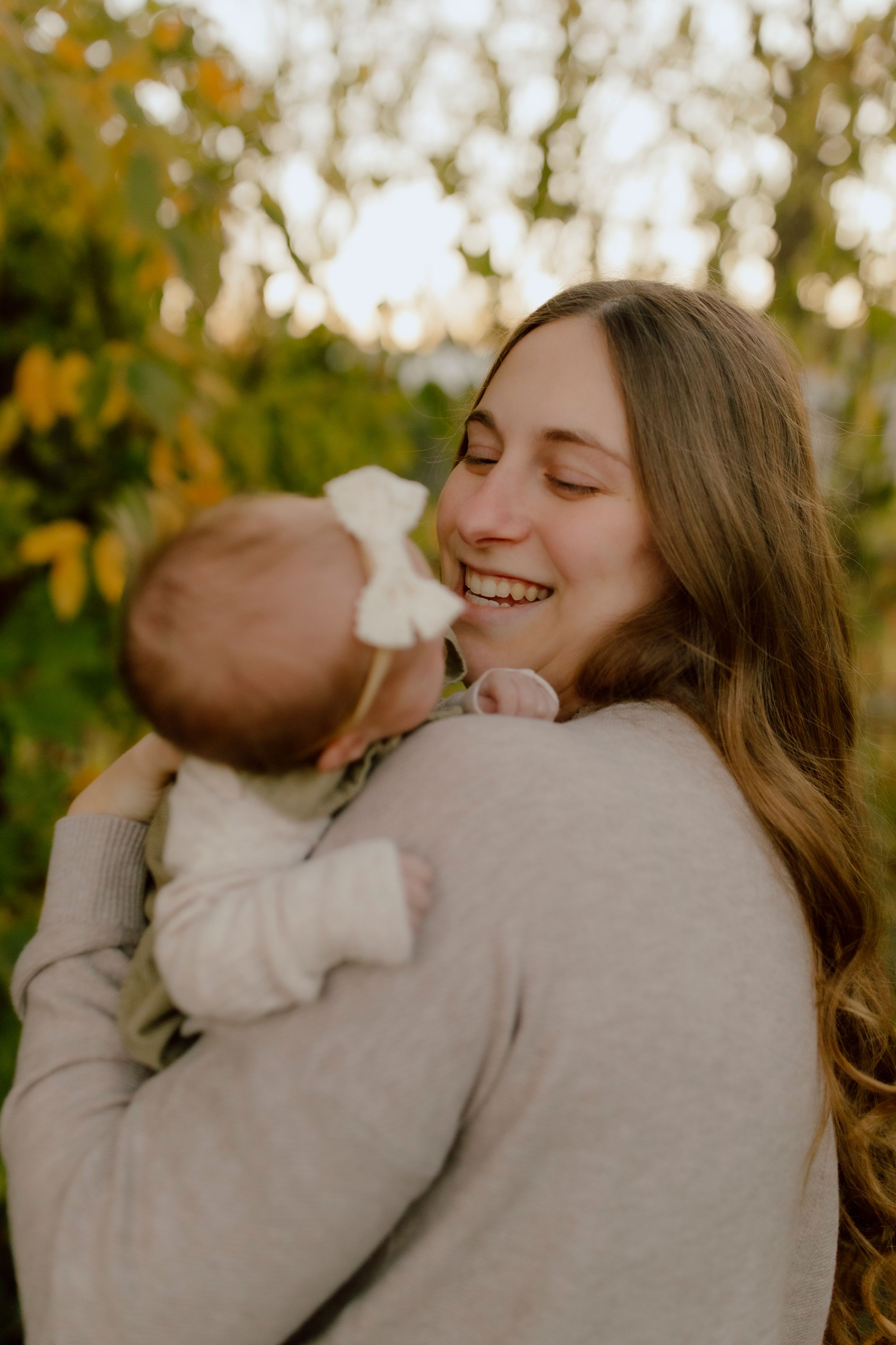 Joyful mother with long hair smiling while holding newborn baby outdoors in autumn setting.