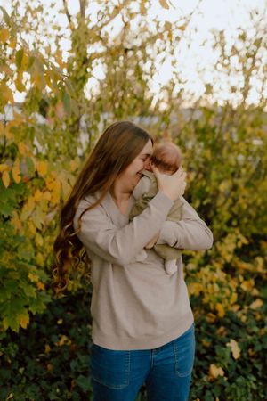 Young mother with long hair cuddling newborn baby outdoors among autumn foliage.