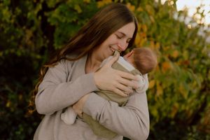 Smiling mother cradles her newborn baby in arms outdoors surrounded by lush green foliage.