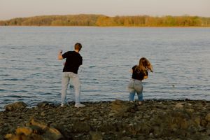 Two people skipping stones along a rocky shoreline at sunset by a calm lake.
