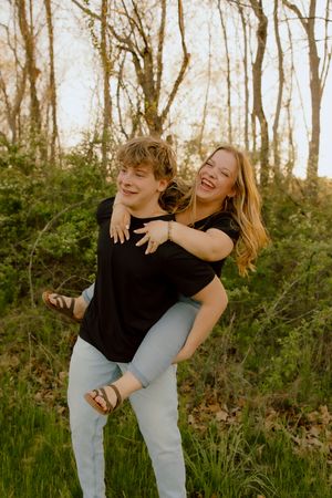 Young couple laughing outdoors, man giving woman piggyback ride in forest setting during golden hour.