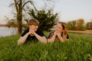 Young couple lying in green grass by lakeside, smiling at each other during sunset.