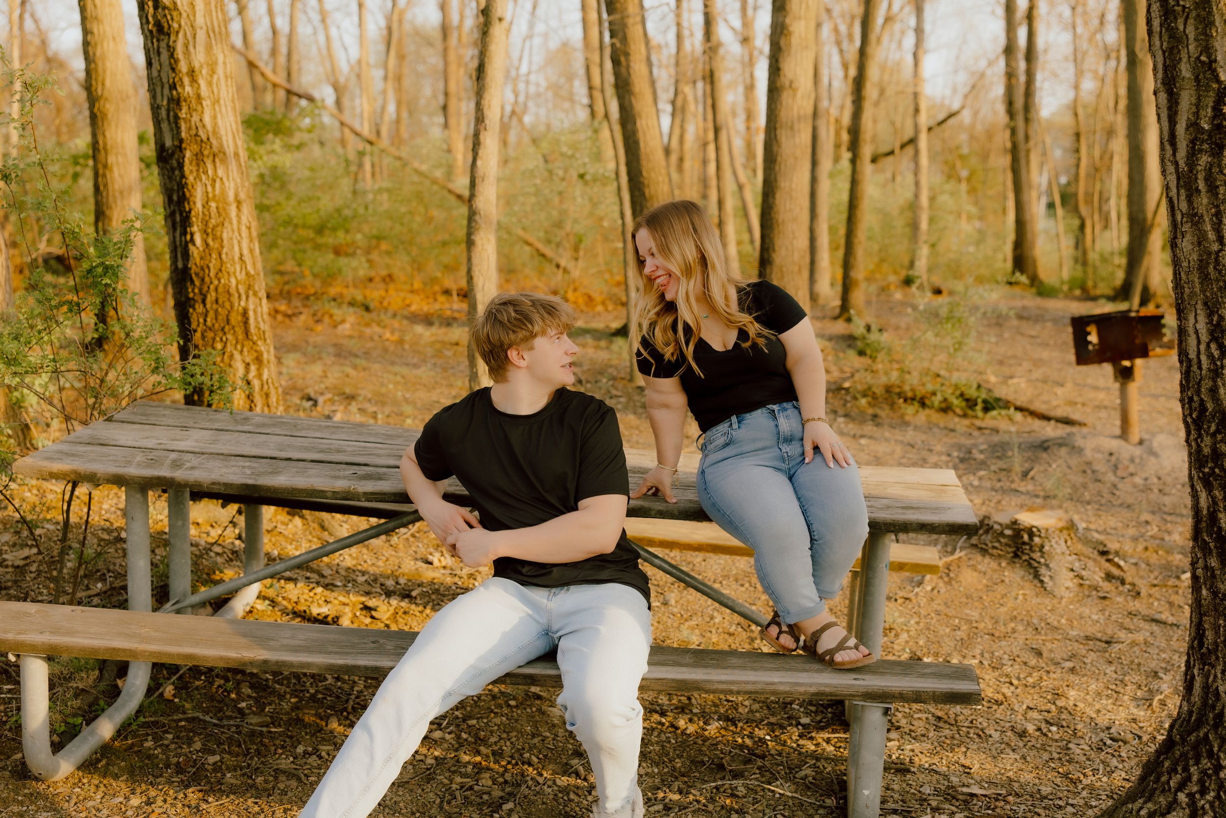 Young couple sitting together on picnic table bench in autumn forest, smiling at each other.