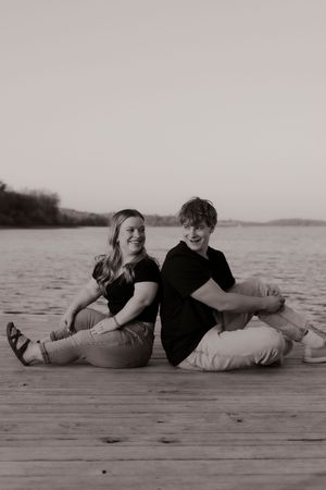 Black and white photo of young couple sitting back-to-back on wooden dock by lake.