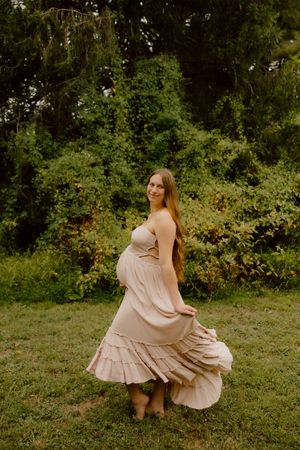 Pregnant woman in flowing beige dress twirling in lush green garden.