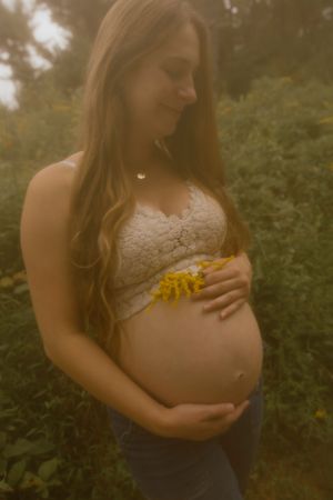 Pregnant woman in lace bralette holding baby bump with yellow wildflowers outdoors.