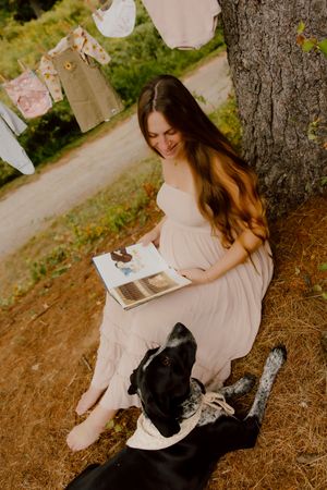 Smiling pregnant woman reads book under tree with black dog beside her.