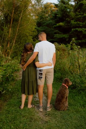 Couple and their brown dog stand on a garden path holding an ultrasound photo announcement.