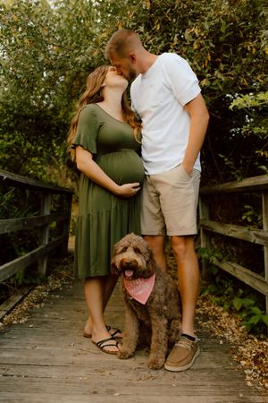 Pregnant couple kissing on a wooden bridge with their fluffy dog wearing a pink bandana.