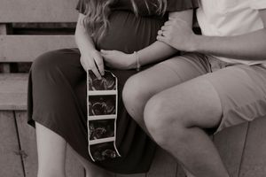 Black and white photo of a pregnant couple sitting together holding ultrasound images.