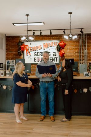 Three people standing and smiling inside Redding Medical Scrubs and Equipment store with balloons.