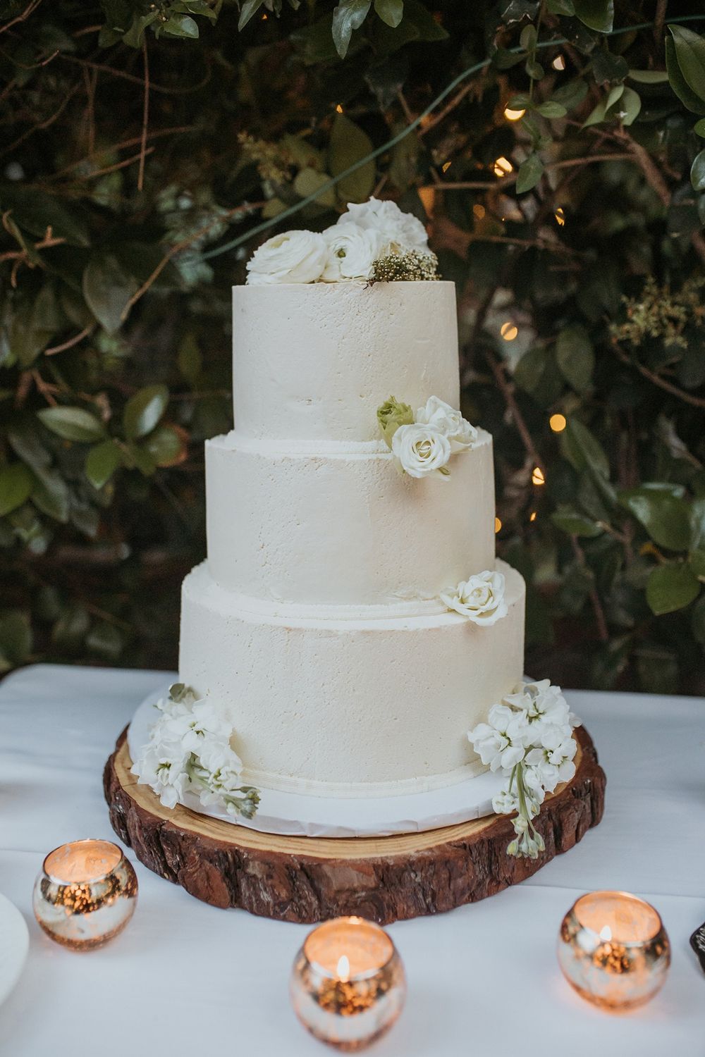 Elegant three-tier white wedding cake decorated with white flowers sits on rustic wood slice with rose gold candles Inn of the Seventh Ray.