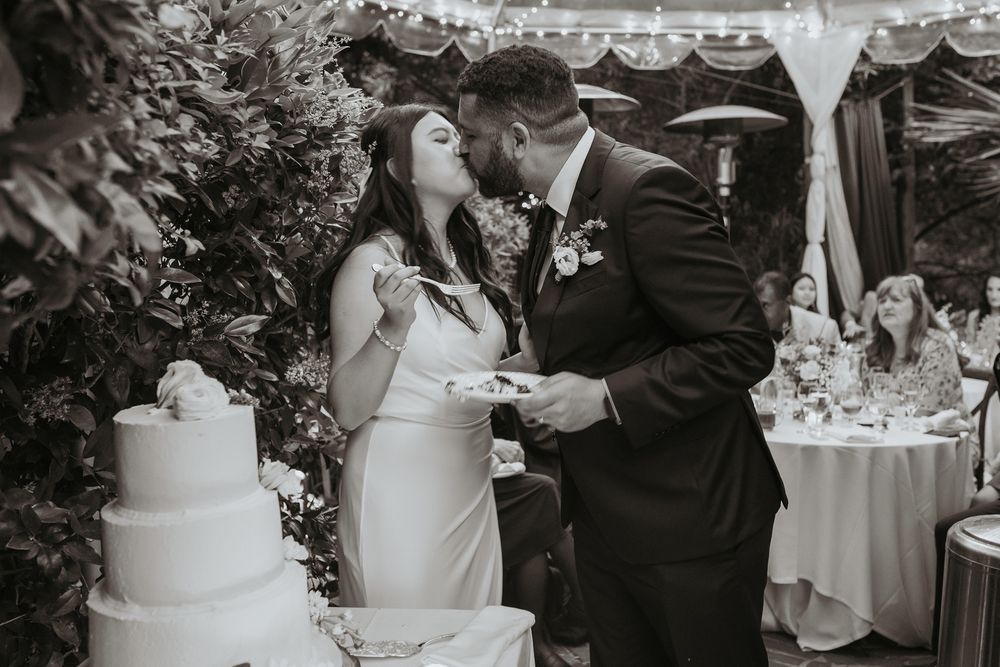 A black and white wedding photo showing a couple sharing a moment while cutting their elegant tiered cake at an outdoor reception.