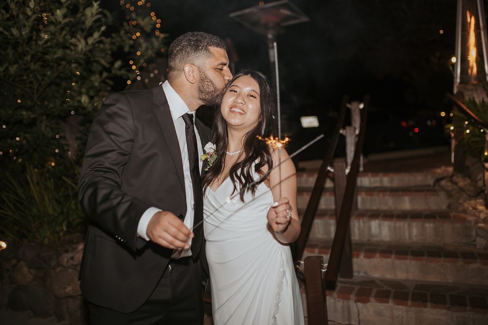 A couple shares a romantic moment with sparklers during their wedding celebration at night with decorative lights in the background.