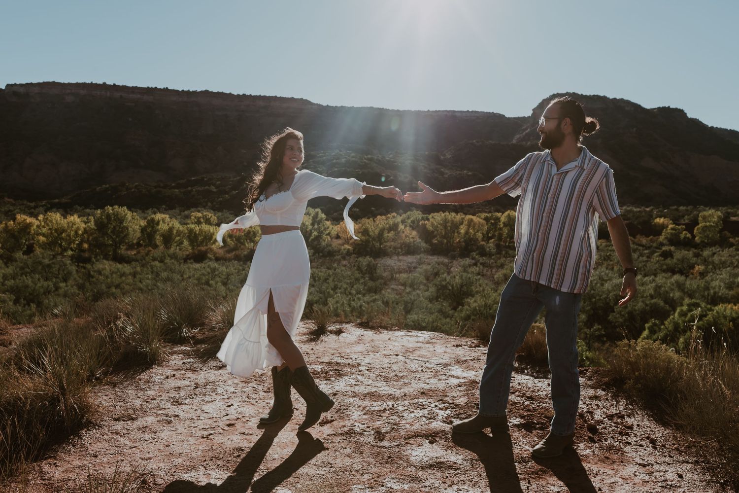 Couple dancing and holding hands on a desert trail with mountains and sunlight in the background.