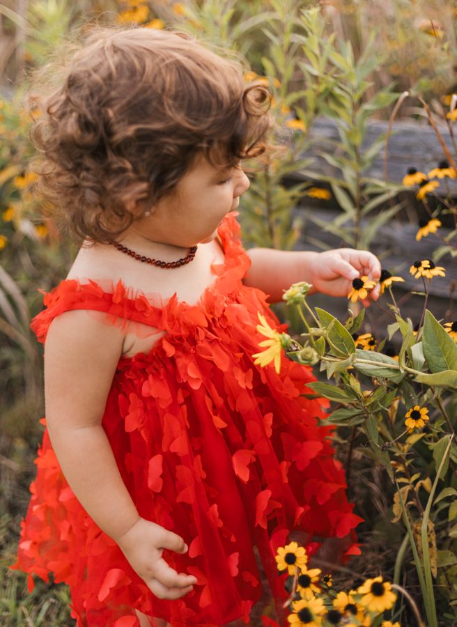 2 year old picky yellow flowers in a red butterfly dress with short curly brown hair with an amber baltic teething necklace on