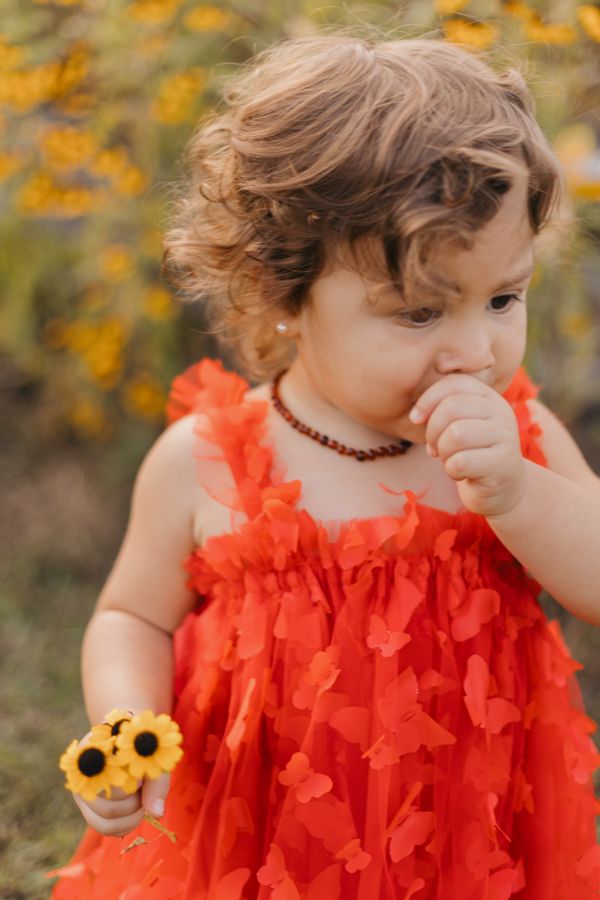 2 year old holding yellow flowers in her hand wearing a red butterfly dress with short curly brown hair while sucking her thumb