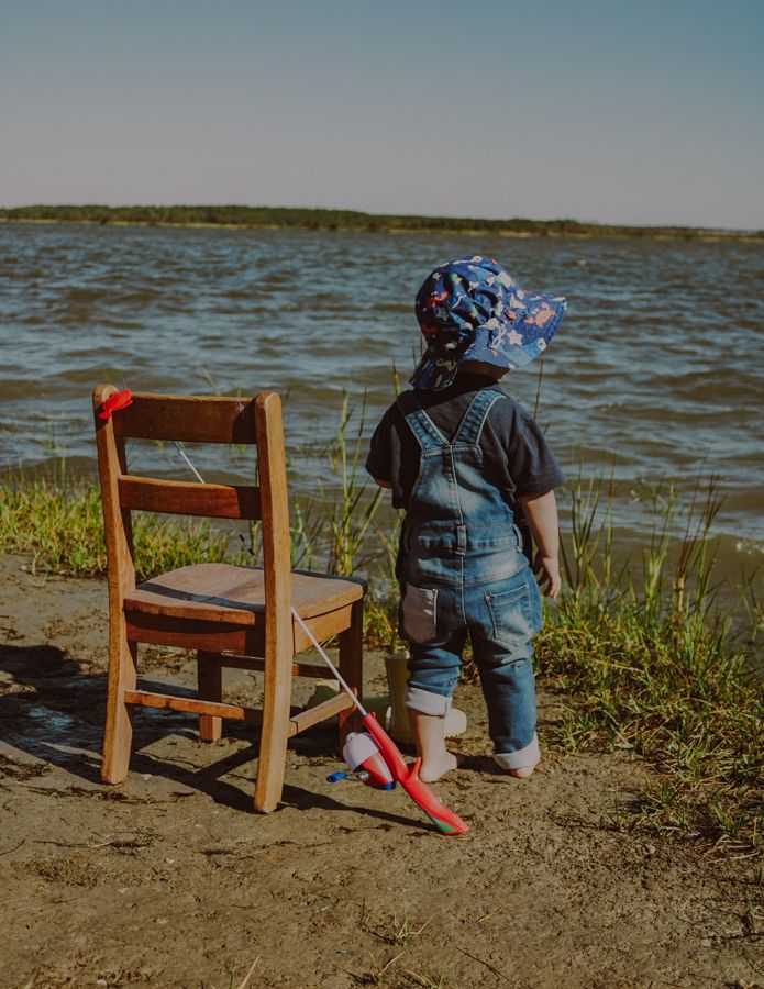 2 year old boy fishing on the York river shore front, in coveralls with a fishing hat with bare feet next to a chair