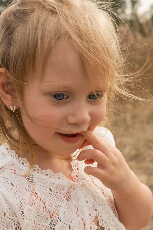 Close-up of blonde toddler girl in white lace dress touching her mouth.