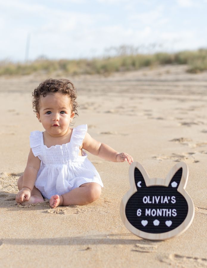 6 month old baby girl  with short curly brown hair on a sandy plain with beach grass in th background