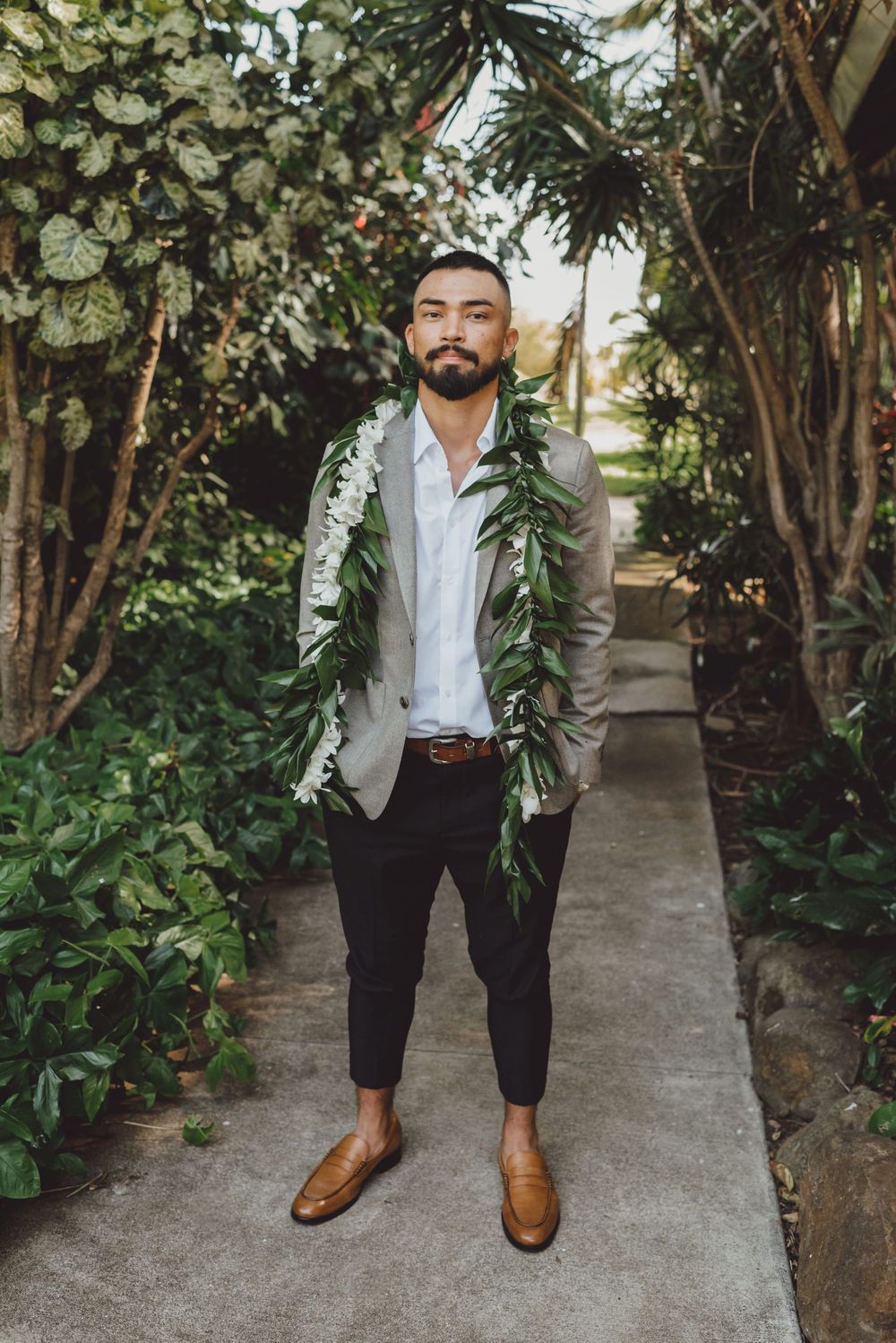 Stylish brown leather dress shoes and black pants shown on a stone pathway surrounded by tropical foliage.