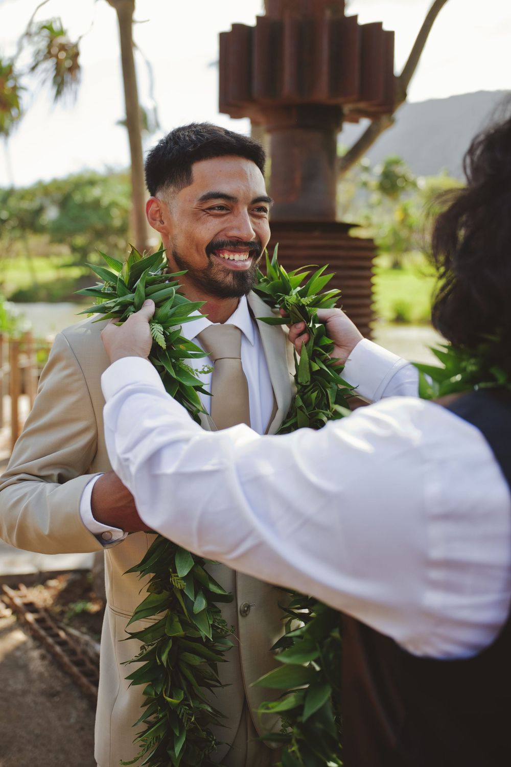 A groomsman wearing a tan suit and white lei receives a traditional Hawaiian maile lei during a wedding ceremony.