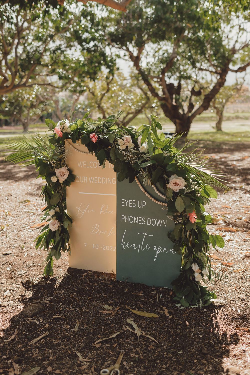 Wedding ceremony sign decorated with lush greenery and flowers displays 'Eyes up phones down' message.
