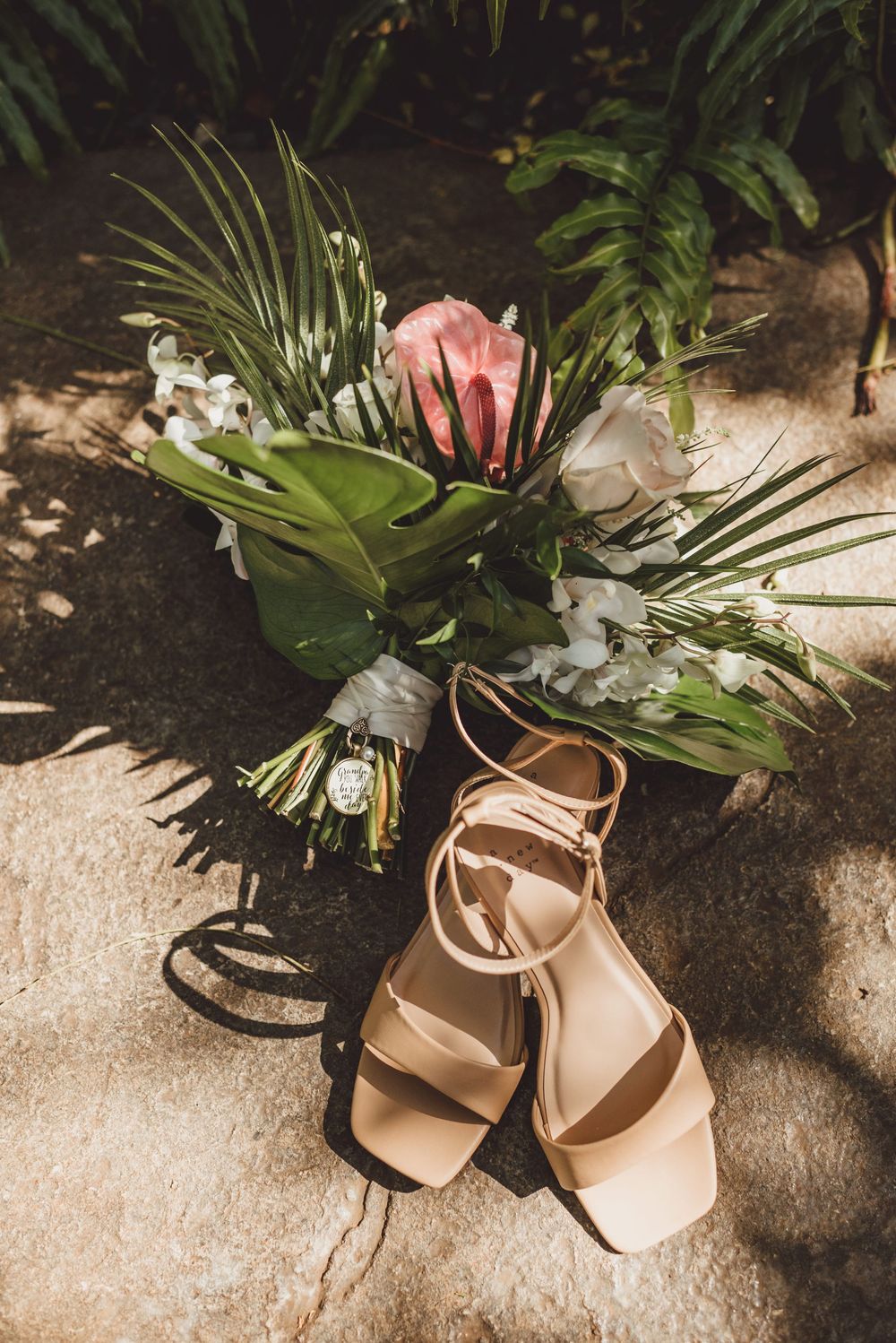 Bridal bouquet with tropical leaves and pink protea flower next to nude wedding sandals.