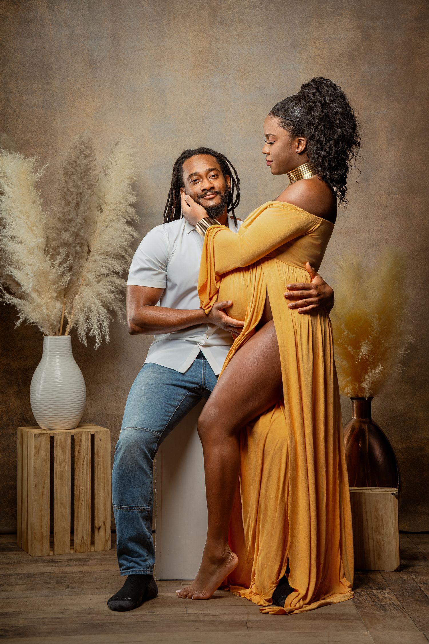Expecting couple poses together in studio with pampas grass decor, woman in flowing yellow dress and man in casual attire.