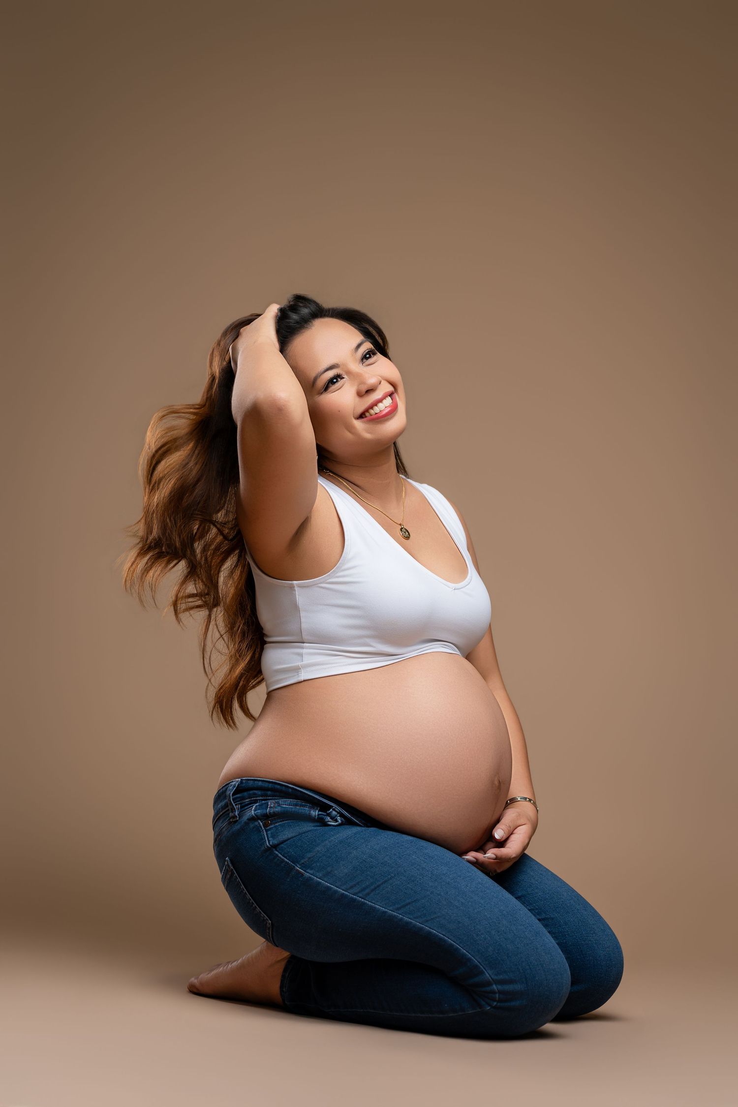 Maternity photoshoot series showing pregnant woman in white top and jeans posing against brown backdrop.