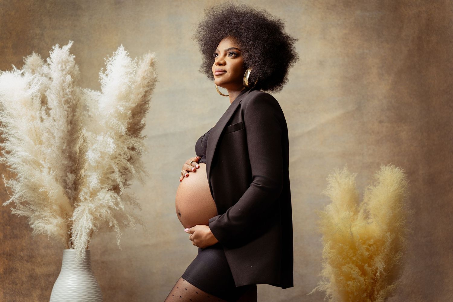 Elegant maternity photo featuring a pregnant woman in black against a beige backdrop with decorative pampas grass arrangements.